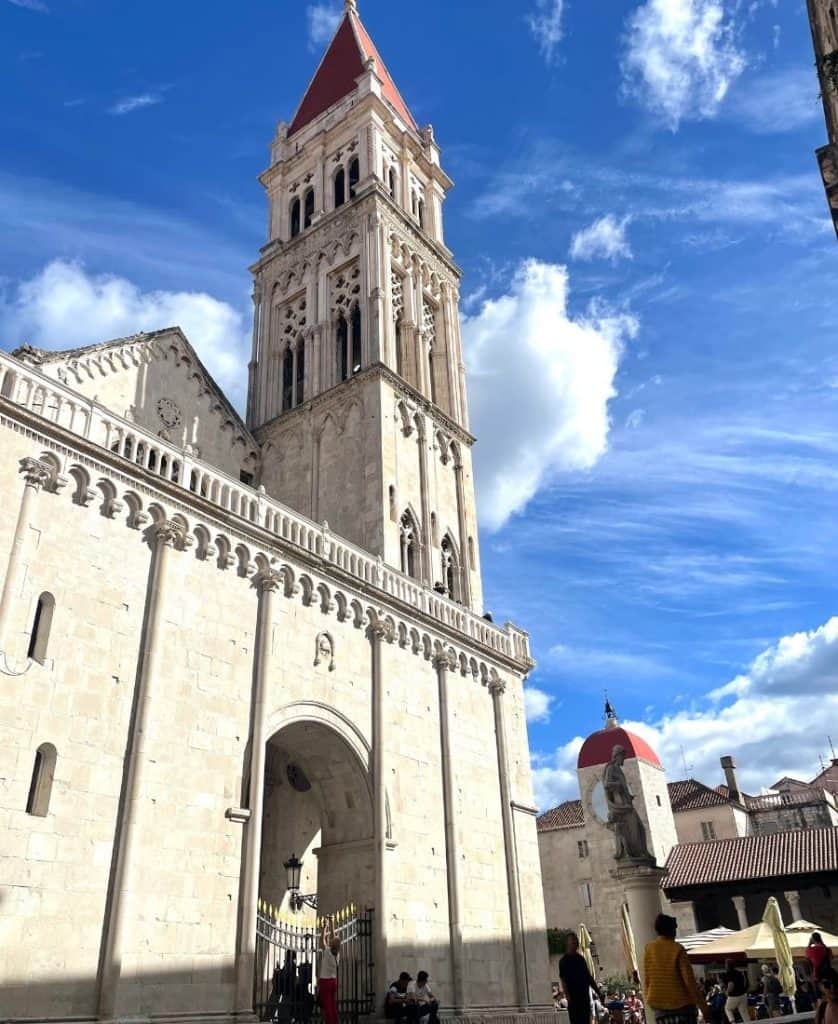 A side view of a triple-naved basilica is pictured with a tall bell tower. It is constructed in a Romanesque-Gothic style. Above it the sky is blue with some clouds.