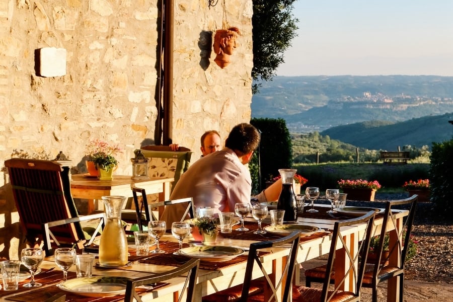 Two people sit at an outdoor dining table set for dinner at an Umbria Agriturismo with a hill town of Italy beyond.
