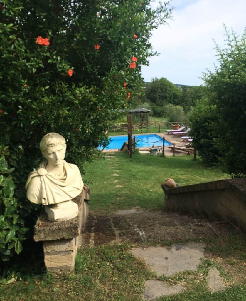 An Italian bust is set next to stone steps that lead to a pool. A shrub with orange flowers is next to the bust.