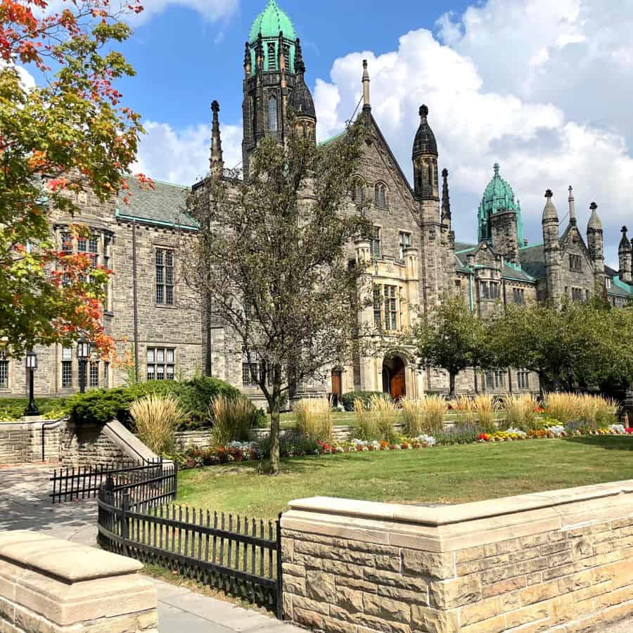 An old building that is part of Trinity College of University of Toronto. Pictured on a fall day with a colorful tree and blue sky.