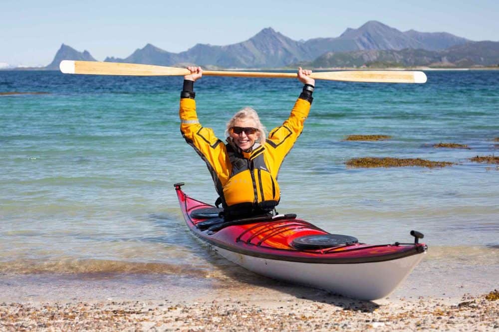 An older woman sits in a kayak holding a paddle in the air. She is at a beach with a blue sea and mountains beyond.