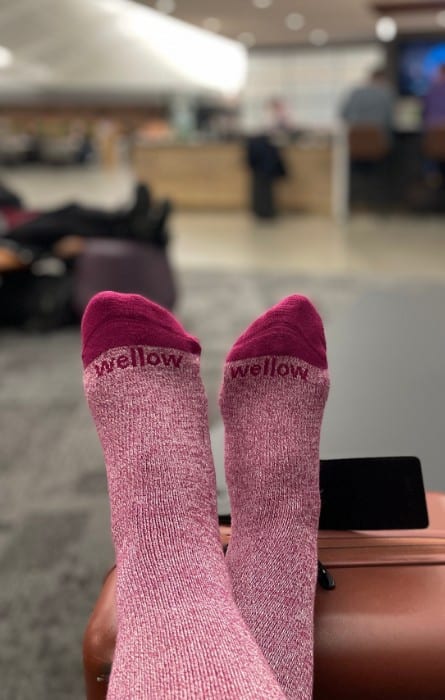 A pair of lower legs and feet are pictured wearing rose colored compression socks. The feet are resting on a carry-on bag in an airport.
