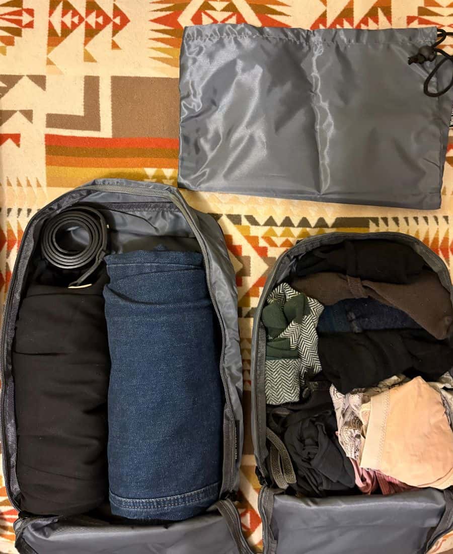 Two packing cubes of different sizes are packed with clothing items. And a laundry bag is pictured above. They are laid flat on a patterned blanket.