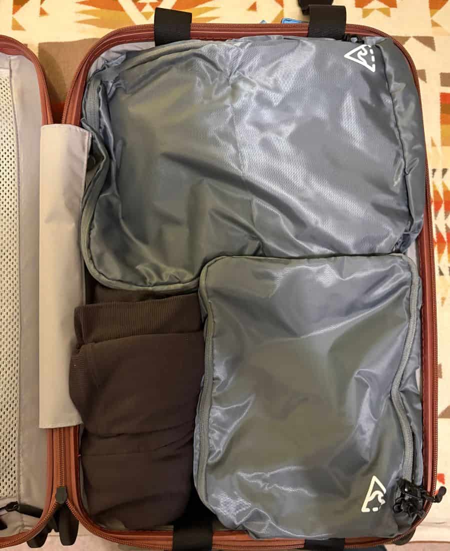 A carry on roller bag is open and packed on one side with two packing cubes and a brown swearer wrap.