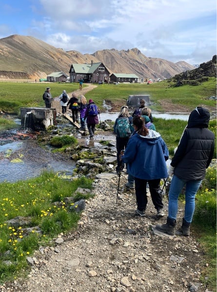 A group of women follow a trail in Iceland. In the distance is a wooden house and some hills.