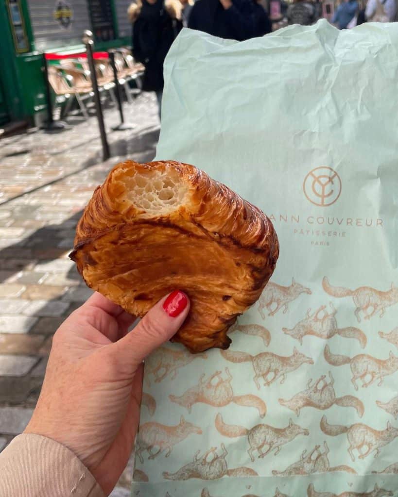 Susan's hand holds a butter croissant and a paper bag of mint green with the words Yann Couvreur. Beyond are cobblestones streets in Paris.