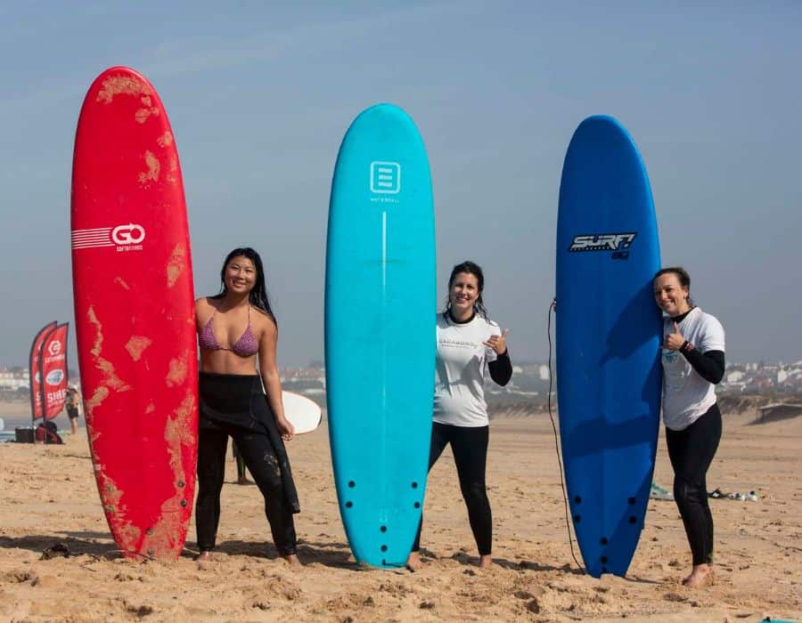Three women stand on the beach with surfboards.