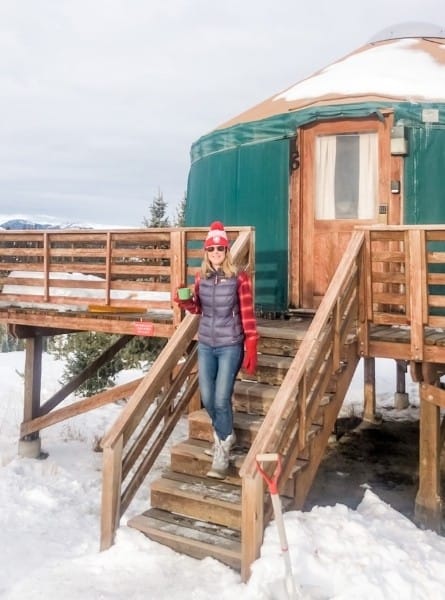 A woman stands on the steps of a yurt surrounded by snow