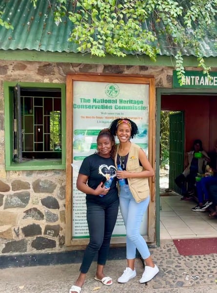 Stella and Tloco, two women guides and travel entrepreneurs, pose together in front of a sign at the entrance to Victoria Falls in Zambia. They are dressed casually in jeans and t shirts and both are smiling.