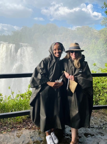 Tloco Kelebemang and Susan Heinrich pose together in rain ponchos, in front of Victoria Falls in Zambia. The falls and a misty spray are visible beyond them, as well as some green trees.