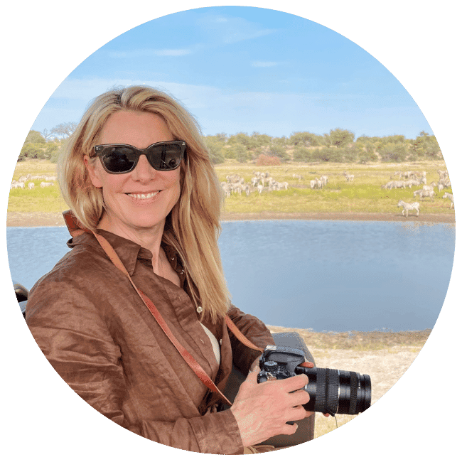 A woman wears a brown linen short and holds a camera. Water and a herd of zebra are just visible in the distance