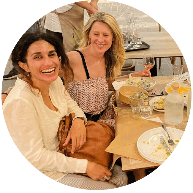 Two women sit together in an outdoor cafe. One is looking at the other and one at the camera. Both are smiling.